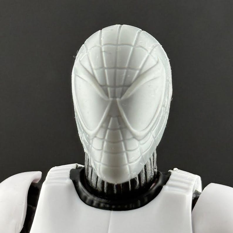 May include: A close-up of a white action figure head with a Spider-Man mask design. The mask has a webbed pattern and large eye openings. The figure's body is white with black accents around the neck.