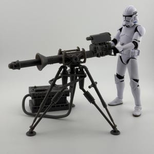 May include: A gray plastic toy model of a Star Wars blaster cannon on a tripod stand. The cannon has a long barrel and a large, detailed engine. A gray plastic toy model of a Star Wars clone trooper stands next to the cannon.