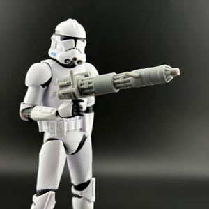 Peut inclure: Figurine blanche d'un Stormtrooper de Star Wars, tenant un blaster gris. La figurine est en position debout, avec un casque et une armure d&eacute;taill&eacute;s. Le blaster est une arme cylindrique &agrave; plusieurs canons.