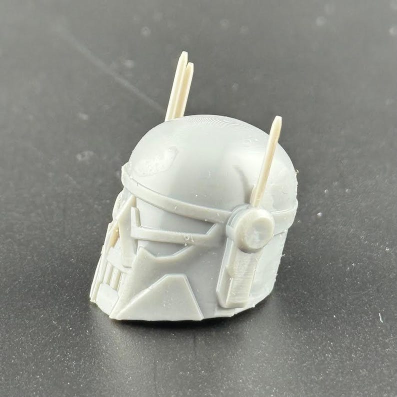 May include: A gray, 3D-printed helmet with a stylized design, featuring a faceplate and two vertical protrusions. The helmet has a smooth, matte finish and appears to be a model or prototype.