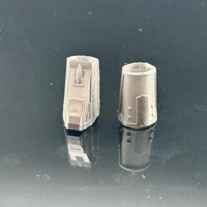 Pu&ograve; includere: Due piccoli oggetti cilindrici grigio metallizzato con dettagliati disegni geometrici. Uno ha una sporgenza centrale verticale. Gli oggetti sono posti su una superficie scura e riflettente, suggerendo uno scatto di prodotto per un componente tecnico o meccanico.