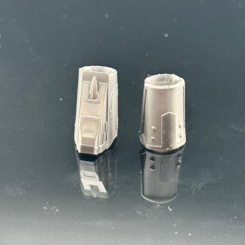 Pu&ograve; includere: Due piccoli oggetti cilindrici grigio metallizzato con dettagliati disegni geometrici. Uno ha una sporgenza centrale verticale. Gli oggetti sono posti su una superficie scura e riflettente, suggerendo uno scatto di prodotto per un componente tecnico o meccanico.