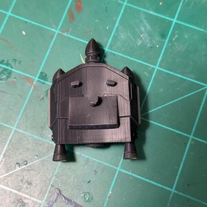 以下が含まれることがあります： 未来的な乗り物または構造物の黒い3Dプリントモデル。モデルは、丸みを帯びた特徴、尖った上部、および基部に小さな円筒形の突起を備えた長方形のボディを持っています。緑色のカッティングマットの上にあります。