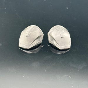 Pu&ograve; includere: Due piccoli oggetti argentati a forma di casco. Ognuno ha una parte superiore arrotondata con un dettaglio rettangolare e una serie di linee. Gli oggetti sono su una superficie riflettente.