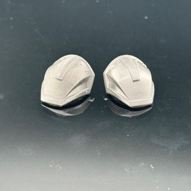 Pu&ograve; includere: Due piccoli oggetti argentati a forma di casco. Ognuno ha una parte superiore arrotondata con un dettaglio rettangolare e una serie di linee. Gli oggetti sono su una superficie riflettente.