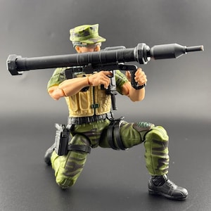 May include: A military action figure kneeling and aiming a black rocket launcher. The figure wears a green camouflage uniform, a tan vest, and a matching cap. The figure is posed as if preparing to fire the weapon.
