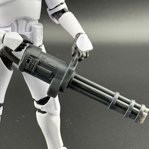 May include: A black and grey miniature minigun held by a white action figure. The minigun features multiple barrels and a detailed design. The action figure is wearing a white suit with black accents.