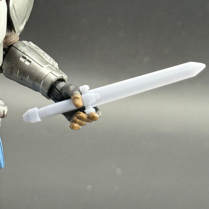 Puede incluir: Una espada de pl&aacute;stico blanco sostenida por una figura con armadura plateada met&aacute;lica. La espada tiene una hoja recta y una empu&ntilde;adura sencilla. Se ve la mano de la figura agarrando firmemente la espada. El fondo es gris neutro.