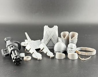 Könnte beinhalten: Eine Sammlung grauer Miniatur-Militäraccessoires, darunter eine Minigun, Pistolen, eine Weste, Stiefel, ein Helm, ein Kopf und ein Gürtel. Diese Accessoires sind wahrscheinlich für Actionfiguren oder Modellbausätze.