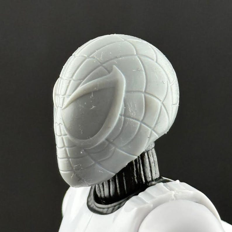 May include: Close-up of a gray Spider-Man action figure head. The mask features a webbed design and large eye openings. The figure's neck is black, and the body is white. The figure is a collectible toy.