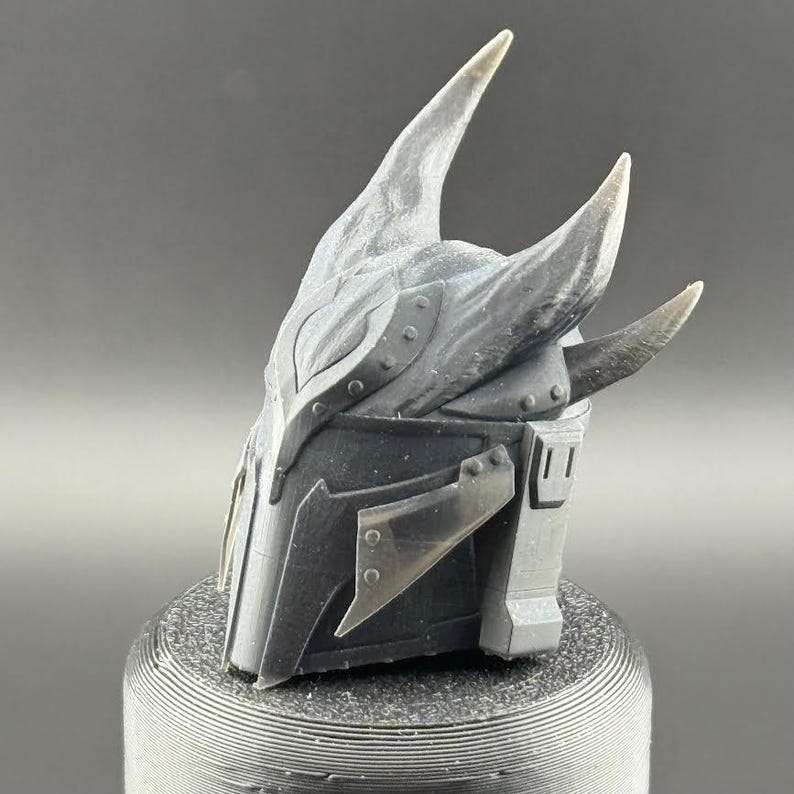 May include: A 3D printed helmet with a dark gray finish and a textured surface. The helmet has two large horns on the top and a smaller horn on the side. The helmet is designed to look like a fantasy or sci-fi character.