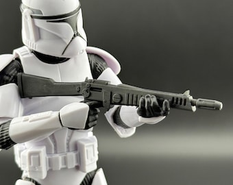 May include: A white and black action figure of a clone trooper holding a black rifle. The trooper is wearing a white helmet and armor with black accents. The figure is posed as if ready for battle.