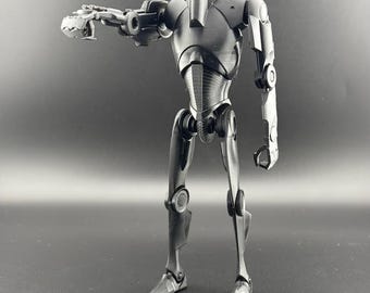 Könnte beinhalten: Eine schwarze und graue, bewegliche Actionfigur eines Roboters mit einem humanoiden Körper und einem großen Kopf. Der Roboter hat einen großen, runden Kopf mit einem einzigen Auge und einem kleinen Mund. Die Arme des Roboters sind lang und dünn, und seine Beine sind kurz und dick. Der Roboter steht auf einer schwarzen Oberfläche.