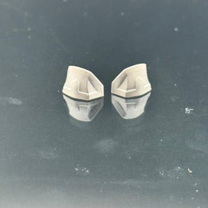 Pu&ograve; includere: Due piccoli oggetti di forma geometrica grigio chiaro con una superficie riflettente. Ogni oggetto ha un design unico sulla superficie superiore, con una serie di linee e angoli. Gli oggetti sono posizionati vicini, con i loro riflessi visibili sotto.