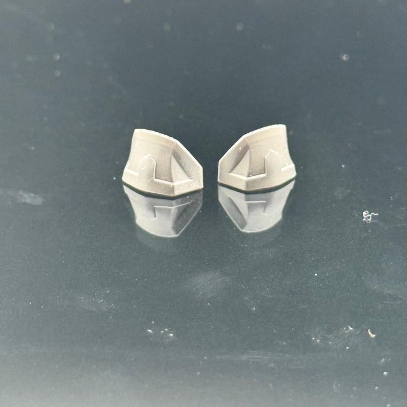 Pu&ograve; includere: Due piccoli oggetti di forma geometrica grigio chiaro con una superficie riflettente. Ogni oggetto ha un design unico sulla superficie superiore, con una serie di linee e angoli. Gli oggetti sono posizionati vicini, con i loro riflessi visibili sotto.