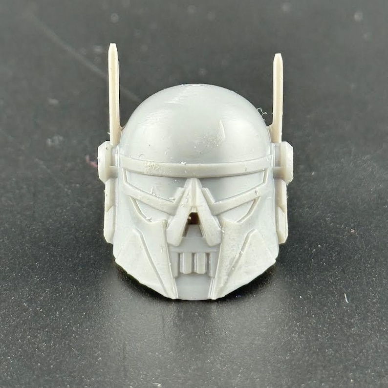 May include: A gray, plastic helmet with a detailed, angular design. The helmet features a visor, a mouth-like opening, and two vertical antenna-like protrusions. The overall aesthetic suggests a futuristic or sci-fi theme.