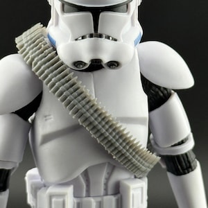 May include: A white action figure of a stormtrooper from Star Wars. The figure has a detailed helmet, armor, and a gray ammunition belt across the chest. The figure's design includes black accents and blue stripes.