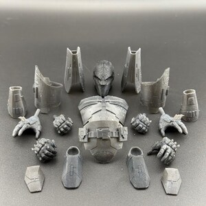 May include: A set of 15 grey plastic action figure parts, including a head, torso, arms, legs, and hands. The parts are arranged on a grey surface.