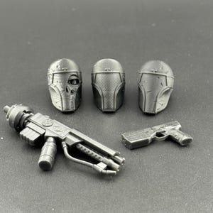 May include: A collection of gray, futuristic-looking toy components, including three helmets, a blaster rifle, and a handgun. The helmets have detailed designs, and the weapons have intricate mechanical features.