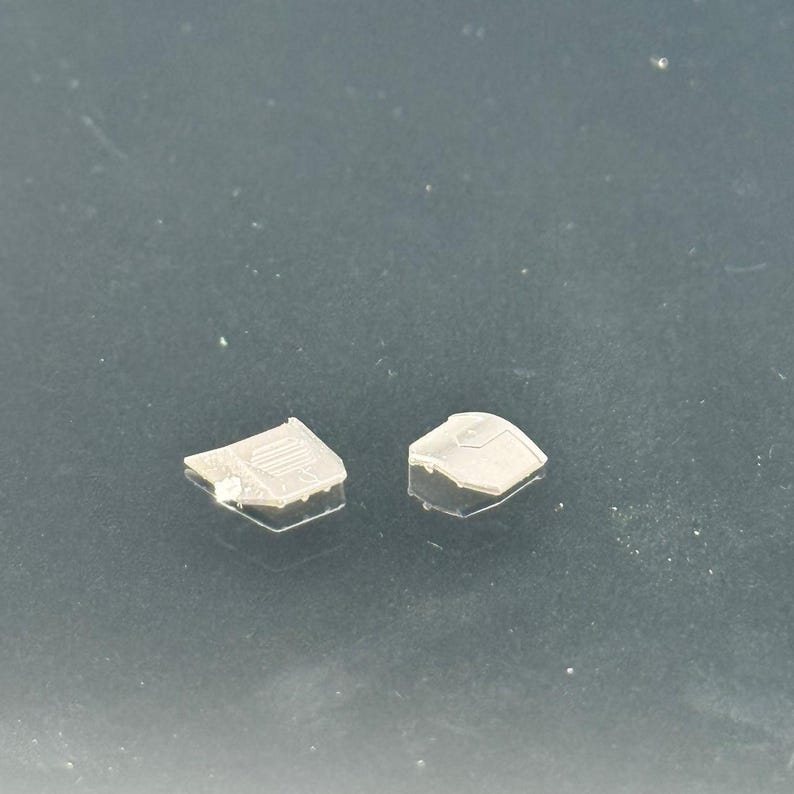 Pu&ograve; includere: Due piccoli pezzi di plastica traslucida con forme geometriche. Un pezzo ha una serie di linee parallele, mentre l'altro ha una parte superiore curva. I pezzi sono su una superficie scura.