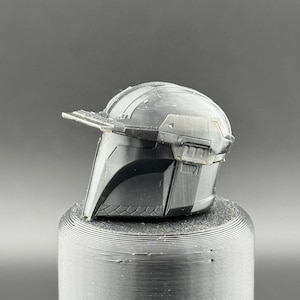Pu&ograve; includere: Un casco di plastica nero con una visiera e una cresta rialzata sulla parte superiore. Il casco &egrave; seduto su una base circolare nera.