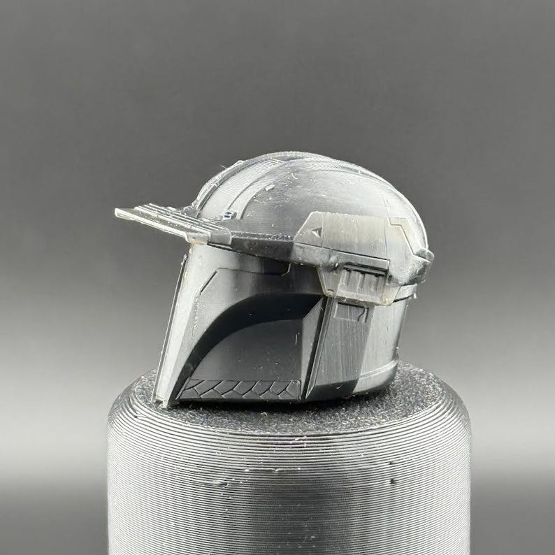 Pu&ograve; includere: Un casco di plastica nero con una visiera e una cresta rialzata sulla parte superiore. Il casco &egrave; seduto su una base circolare nera.
