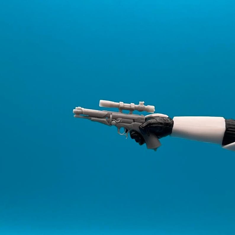 May include: A gray plastic toy blaster with a black scope and a black handle. The blaster is held in a hand.