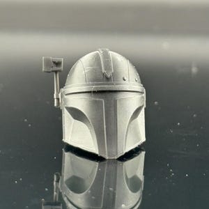 Pu&ograve; includere: Un casco grigio Mandaloriano, una replica dell'iconico copricapo. Il casco presenta una visiera a forma di T e un telemetro sul lato destro. La superficie ha una finitura opaca e il design &egrave; dettagliato.