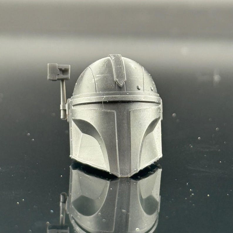 Pu&ograve; includere: Un casco grigio Mandaloriano, una replica dell'iconico copricapo. Il casco presenta una visiera a forma di T e un telemetro sul lato destro. La superficie ha una finitura opaca e il design &egrave; dettagliato.