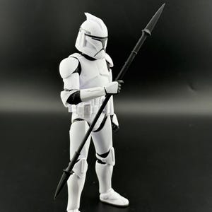 K&ouml;nnte beinhalten: Eine wei&szlig;e Actionfigur eines Klonkriegers aus einem Science-Fiction-Franchise, die einen schwarzen Speer h&auml;lt. Die Figur tr&auml;gt eine wei&szlig;e R&uuml;stung mit schwarzen Akzenten und einen Helm. Der Hintergrund ist einfarbig schwarz.