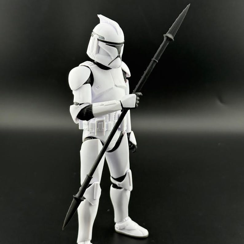K&ouml;nnte beinhalten: Eine wei&szlig;e Actionfigur eines Klonkriegers aus einem Science-Fiction-Franchise, die einen schwarzen Speer h&auml;lt. Die Figur tr&auml;gt eine wei&szlig;e R&uuml;stung mit schwarzen Akzenten und einen Helm. Der Hintergrund ist einfarbig schwarz.