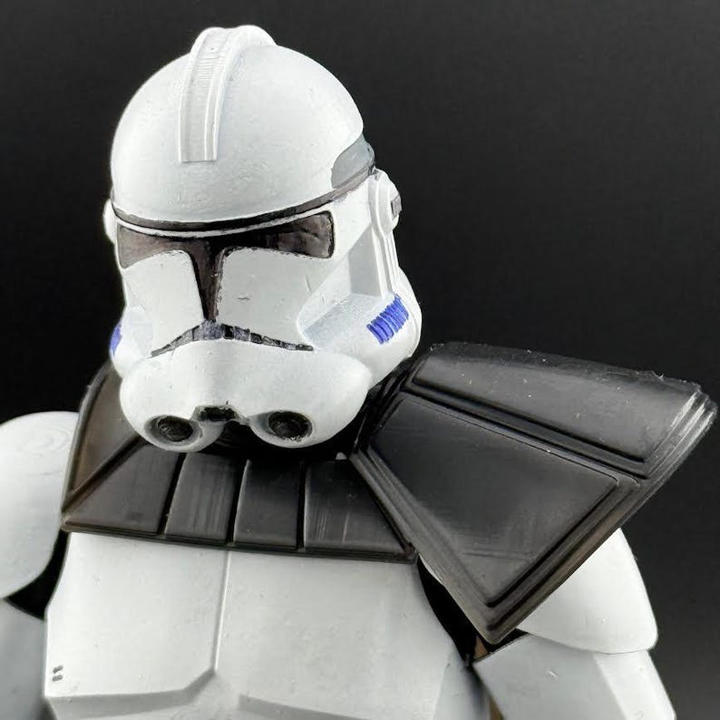 May include: Close-up of a white and black action figure helmet with blue accents. The helmet has a distinctive design with a visor and a central ridge. The figure also has black shoulder armor and a white torso.