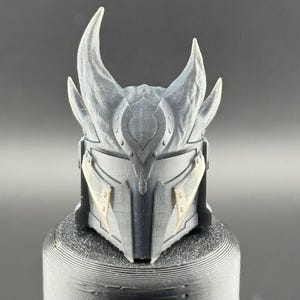 May include: A black and gray 3D printed helmet with horns and decorative details. The helmet has a textured surface and appears to be made of plastic.