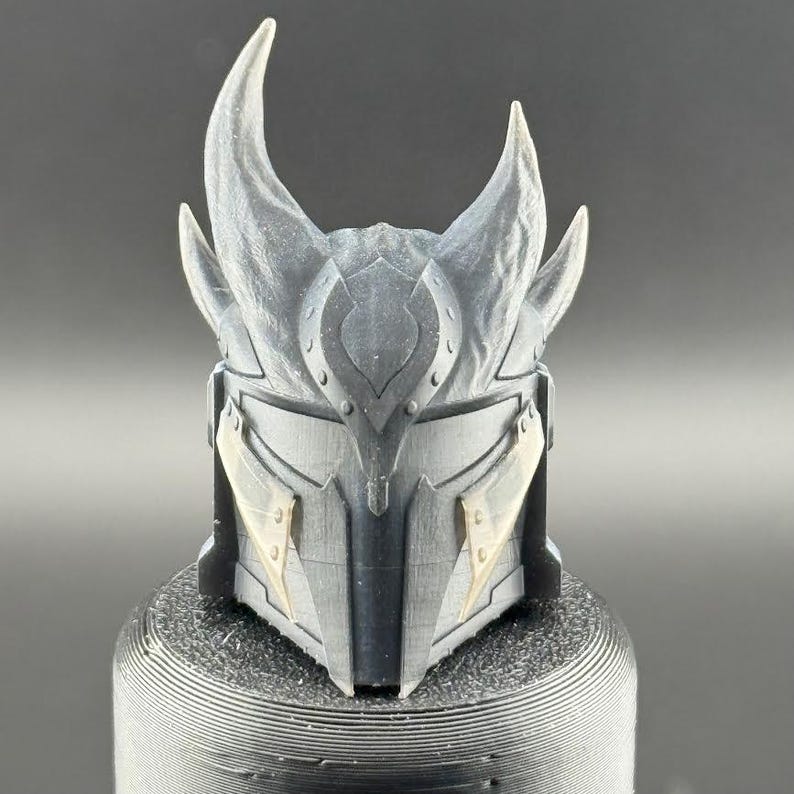 May include: A black and gray 3D printed helmet with horns and decorative details. The helmet has a textured surface and appears to be made of plastic.
