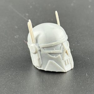 May include: A gray, 3D-printed helmet with a detailed design. The helmet features a visor, cheek guards, and two vertical antenna-like protrusions. The overall design suggests a futuristic or sci-fi theme.