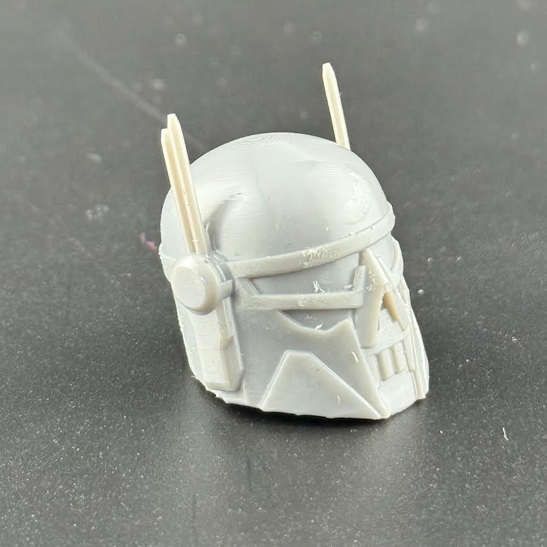 May include: A gray, 3D-printed helmet with a detailed design. The helmet features a visor, cheek guards, and two vertical antenna-like protrusions. The overall design suggests a futuristic or sci-fi theme.