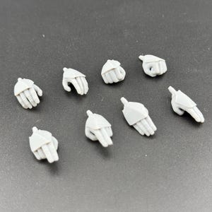 Peut inclure: Un ensemble de huit petites mains accessoires en plastique gris clair non peint pour figurines. Chaque pièce présente une pose distincte, incluant des paumes ouvertes, des poings fermés et diverses positions de doigts, avec une articulation.