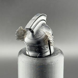 May include: A silver-colored, detailed, helmet with a winged design. The helmet is sitting on a textured, gray surface.