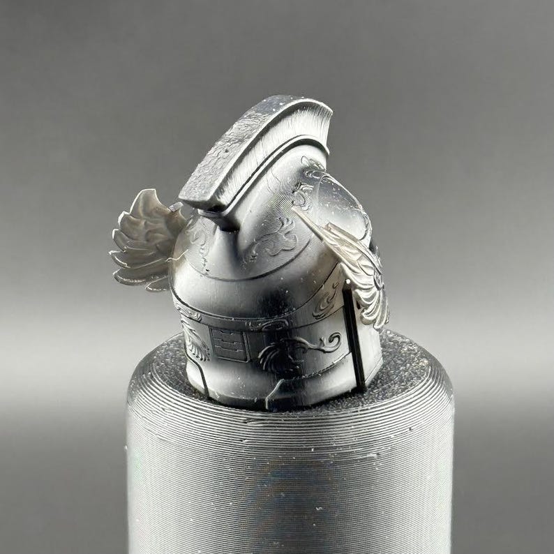 May include: A silver-colored, detailed, helmet with a winged design. The helmet is sitting on a textured, gray surface.