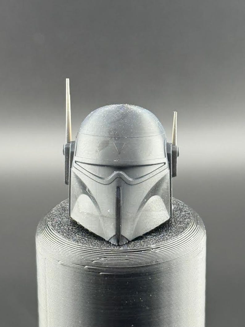 May include: A black helmet with a visor and two antennas. The helmet has a textured surface and a V-shaped design on the front.