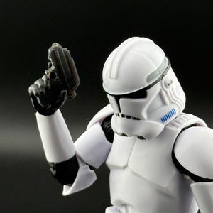 May include: A white action figure of a clone trooper from Star Wars, holding a black blaster pistol. The trooper is wearing white armor with a helmet and black accents. The figure is posed against a dark background.