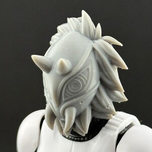 May include: A close-up of a gray, horned helmet with intricate eye-like designs and a spiky, textured crown. The helmet is part of a white Stormtrooper-style armor, suggesting a sci-fi or cosplay theme.