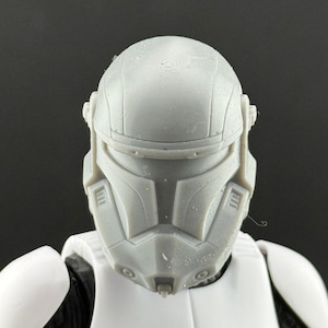 May include: A close-up of a gray, plastic helmet, featuring a detailed design with angular shapes and a visor. The helmet is part of a larger action figure, with white armor visible below. The overall aesthetic suggests a science fiction or military theme.