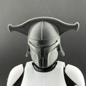 K&ouml;nnte beinhalten: Schwarz-wei&szlig;e Actionfigur einer Figur mit einem Helm mit einem markanten, gebogenen Visier. Der Helm hat ein segmentiertes Design und bedeckt das Gesicht, mit einem wei&szlig;en Brustpanzer und Schulterpolstern. Der Artikel ist ein Sammlerst&uuml;ck.