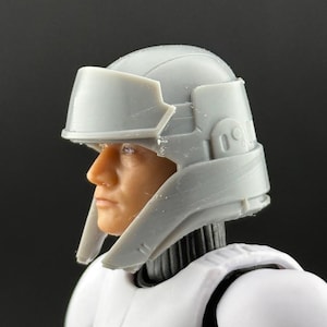 May include: A gray helmet with a visor, part of a white uniform. The helmet has a detailed design with a rounded top and angular visor. The figure's face is visible, with a neutral expression. The overall theme is science fiction.