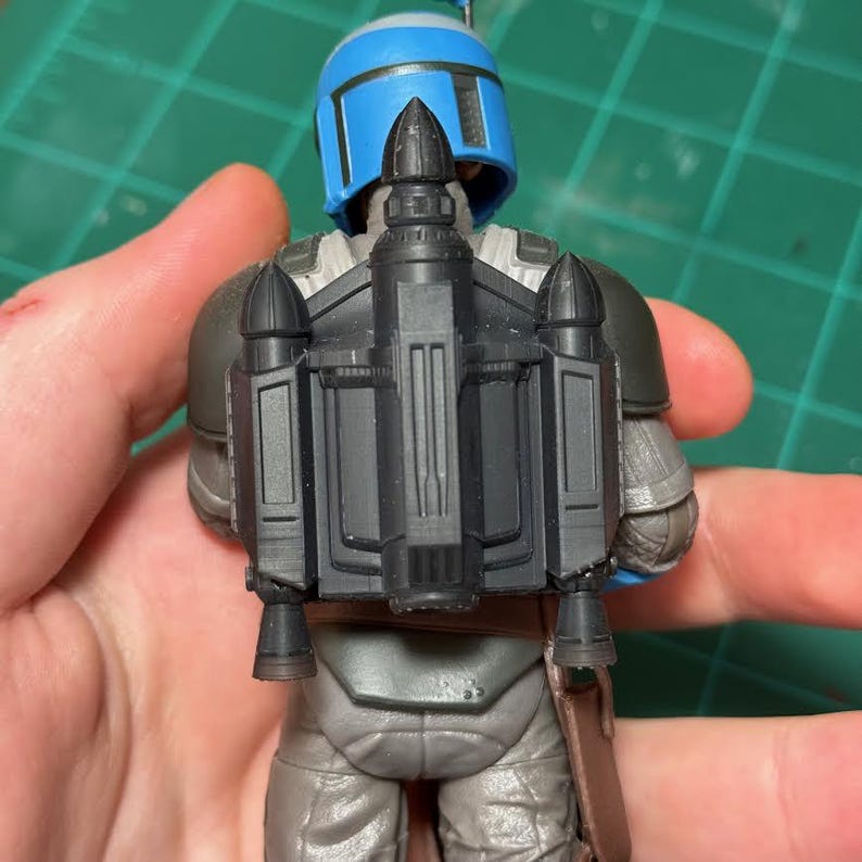 以下が含まれることがあります： SFフランチャイズのキャラクターのアクションフィギュア。青いヘルメット、グレーのアーマー、黒いジェットパックが特徴です。フィギュアは人の手で持たれています。