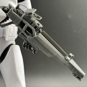 May include: A gray plastic toy blaster rifle held by a white armored action figure. The weapon features a long barrel, a central cylindrical chamber, and a top-mounted sight mechanism. The design includes angular structural supports and a grip.