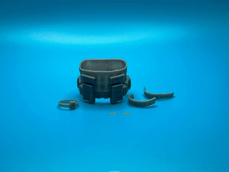 Peut inclure: Une ceinture tactique grise avec plusieurs poches et sangles. La ceinture est d&eacute;mont&eacute;e et dispos&eacute;e sur une surface bleue.