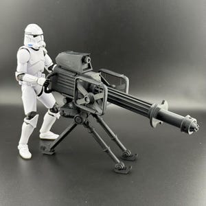 以下が含まれることがあります： 白いストームトルーパーのアクションフィギュアが、黒い重機関銃の隣に立っています。大砲は三脚に取り付けられており、詳細な銃口を備えた大きな銃身があります。フィギュアは、アクションの準備ができた、クラシックなポーズをとっています。