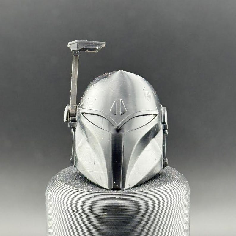 K&ouml;nnte beinhalten: Ein silberner Mandalorianer-Helm mit einem T-f&ouml;rmigen Visier und einem Entfernungsmesser. Der Helm mit seinem kantigen Design steht auf einem strukturierten, zylindrischen Sockel. Das Design ist von der Star Wars-Franchise inspiriert.